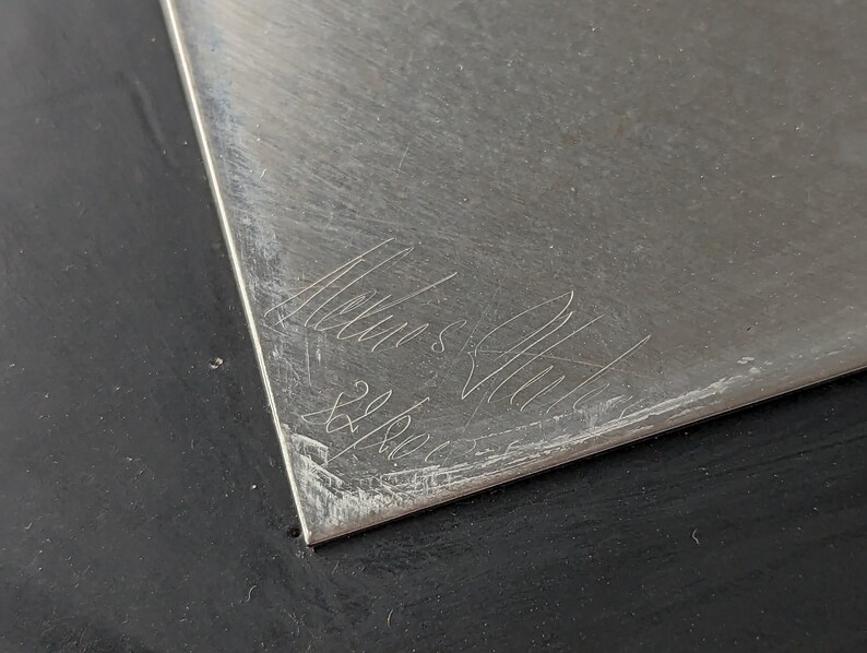 May include: A silver-colored, rectangular metal plate with etched cursive text and the numbers "22/8001". The plate has a brushed texture and is set against a black surface.