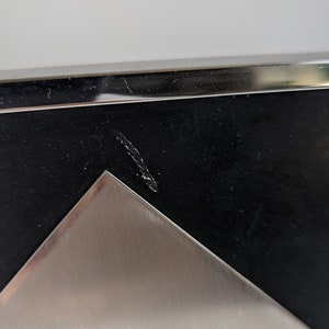 May include: Close-up of a modern picture frame with a black interior and a silver metal corner. The frame has a sleek, minimalist design, with a polished metal edge. The frame is likely for displaying photographs or artwork.