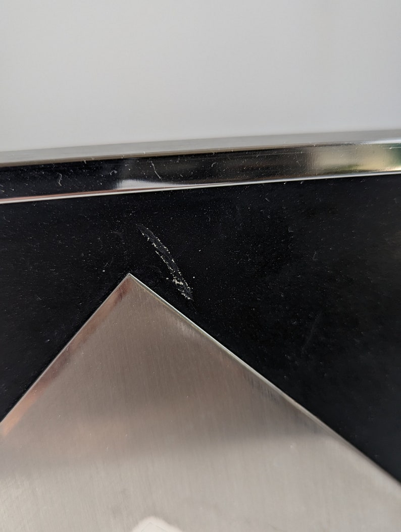 May include: Close-up of a modern picture frame with a black interior and a silver metal corner. The frame has a sleek, minimalist design, with a polished metal edge. The frame is likely for displaying photographs or artwork.