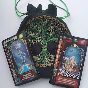 May include: Two tarot cards, The Star and The Hierophant, are displayed on a black fabric bag with a green embroidered tree. The Star card features a woman in a blue gown standing on a globe, with a star above her head. The Hierophant card shows a man in a white robe holding a crystal ball, with a bat flying above him. The text on the cards reads "The Light of the Evening Star shines through Galadriel's King." and "Saruman courts dark and secret forces. The Palantir has given him a vision of his own future glory."