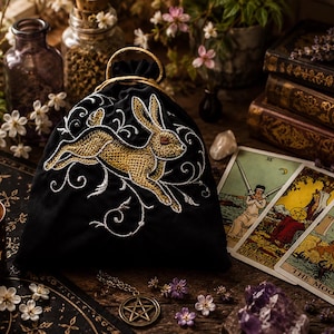 May include: Black velvet drawstring bag with a gold embroidered rabbit design and white accents. The bag is next to tarot cards, a lit candle, and other mystical items. The theme is spiritual and magical.