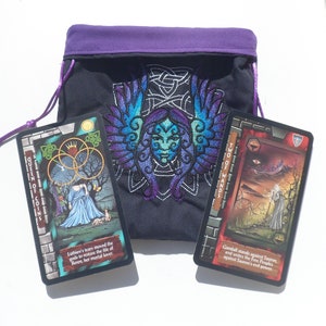 May include: Two tarot cards, Queen of Coins and Two of Wands, are displayed on a black fabric bag with purple trim and an embroidered design. The Queen of Coins card features a woman in a blue dress with a crown and a tree in the background. The Two of Wands card features a man standing on a hill with a staff and a sunset in the background. The text on the cards reads "Queen of Coins" and "Two of Wands".