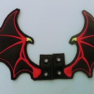 Może przedstawiać: Para czarnych i czerwonych skrzydeł diabła z żółtymi akcentami. Skrzydła są przymocowane do czarnego paska z czterema metalowymi oczkami.