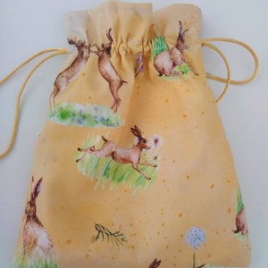 May include: A yellow drawstring bag with a repeating pattern of brown rabbits and a white flower. The bag is made of a soft, fabric material.