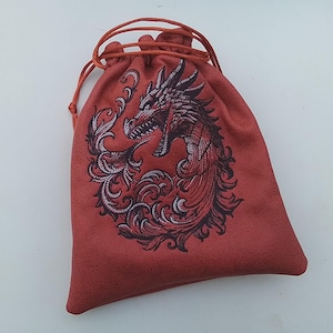 Op de afbeelding: Rode trekkoordtas met een gedetailleerd drakenontwerp in zwart en rood. De draak is omgeven door sierlijke, wervelende patronen. De tas is gemaakt van een textuurrijk materiaal en heeft een rood trekkoord.