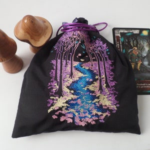 Peut inclure: Un sac en velours noir avec cordon de serrage, orné d'un motif brodé représentant une scène de forêt avec une rivière qui la traverse. La broderie est réalisée dans des tons de rose, bleu et jaune. Le sac est posé sur une surface blanche à côté de deux figurines de champignons en bois.