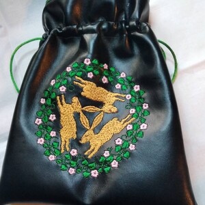 May include: Black leather drawstring bag with a green top and an embroidered design of three rabbits in a circle of green leaves and pink flowers.