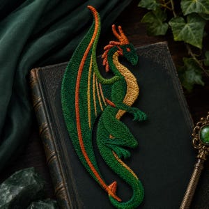 May include: An embroidered patch featuring a green dragon with orange accents on its wings, head, and tail. The dragon is depicted in a side profile with a long, curved tail, a textured underbelly, and detailed wing structures in shades of green and orange.