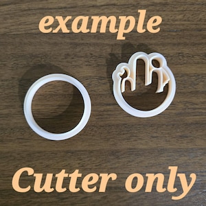 Op de afbeelding: Twee witte plastic koekjesvormpjes. De ene is een eenvoudige ronde ring, de andere is een ronde uitsteker met een intern ontwerp van een gestileerd figuur met een opgeheven hand. De tekst "example" en "Cutter only" zijn in de afbeelding zichtbaar.