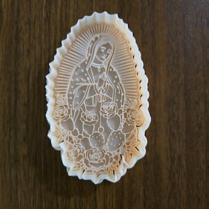 May include: An oval-shaped white cookie cutter featuring an embossed design of a figure with hands clasped in prayer, surrounded by a sunburst halo, roses, and prickly pear cactus pads. The cutter has a scalloped edge and a detailed interior.
