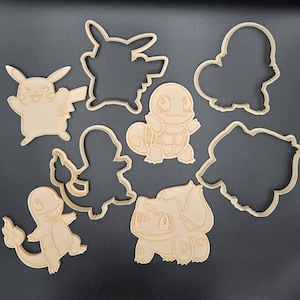 May include: A collection of light brown cookie cutters in the shapes of various cartoon characters, including Pikachu, Squirtle, Charmander, and Bulbasaur. The cutters are arranged on a dark gray surface, showcasing their detailed designs.