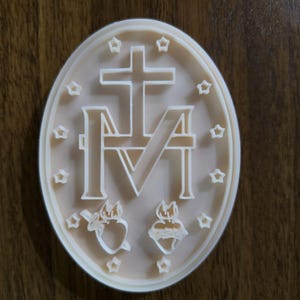 May include: An oval-shaped beige plaque featuring a raised relief design of the Miraculous Medal symbol. The center displays a large letter M surmounted by a cross, flanked by two hearts at the bottom, all encircled by twelve small five-pointed stars.