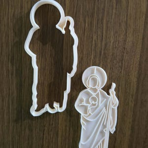May include: Two white plastic cookie cutters in the shape of a religious figure. One is a solid figure, and the other is an outline. The cutters are on a brown wooden surface.