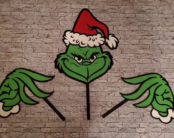 Op de afbeelding: Een groen Grinch-gezicht met een rode kerstmuts en twee groene handen met witte randen. Het gezicht van de Grinch heeft een boze uitdrukking. De handen en het gezicht zijn bevestigd aan zwarte stokken, voor tuindecoratie.
