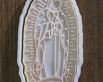 Op de afbeelding: Een beige en witte Maria-koekjesvorm. Het ontwerp toont een gedetailleerde omtrek van de Maagd Maria met lichtstralen die van achter haar uitstralen. De koekjesvorm is ovaal van vorm.