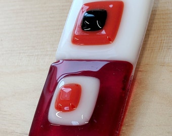 May include: A rectangular pendant with a silver bail. The pendant is composed of fused glass in white, red, and black. The design features layered squares of color, with a black square at the center of the top section.