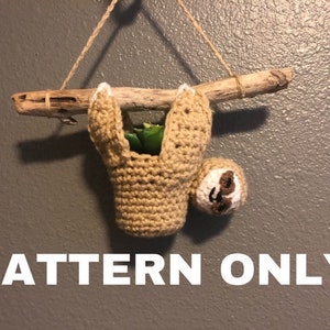 Puede incluir: Un macetero de perezoso tejido a ganchillo en color beige, colgado de una rama de madera flotante con hilo. El perezoso tiene una cara blanca y detalles marrones. La planta es verde. El texto "PATTERN ONLY" se muestra en blanco.