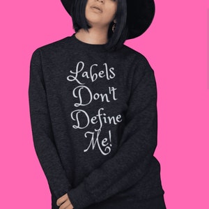 May include: A black sweatshirt with the text "Labels Don't Define Me!" in white lettering. The model is wearing a black hat and black pants.