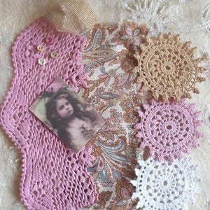 May include: A selection of vintage craft items. It features a pink crocheted doily with buttons, a paisley fabric piece, a small hessian sack, a vintage photograph, and several crocheted doilies in white, pink, and beige.