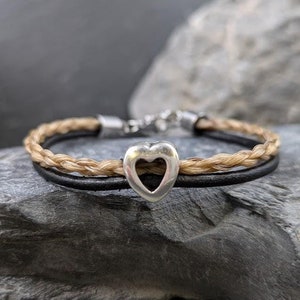 Op de afbeelding: Een gevlochten leren armband met een zilveren hartbedel. De armband is gemaakt van twee leren strengen, een bruine en een beige, en is samen gevlochten. De hartbedel is zilverkleurig en heeft een uitgesneden ontwerp.