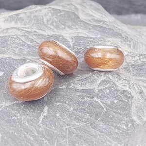 May include: Three brown and silver beads with a natural wood grain appearance. The beads are in different shapes: a barrel, an oval, and a round bead. The beads are set against a gray, textured stone surface.
