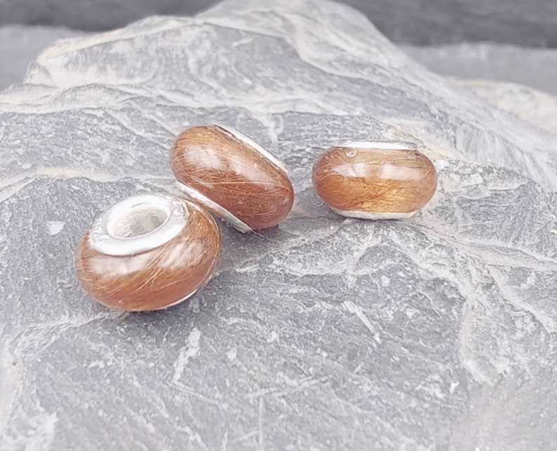 May include: Three brown and silver beads with a natural wood grain appearance. The beads are in different shapes: a barrel, an oval, and a round bead. The beads are set against a gray, textured stone surface.
