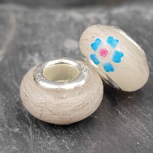 May include: Two decorative beads with silver-colored accents. One bead is round, the other is oval-shaped. The oval bead has a floral design with blue and pink accents. Both beads have a light-colored, textured surface.