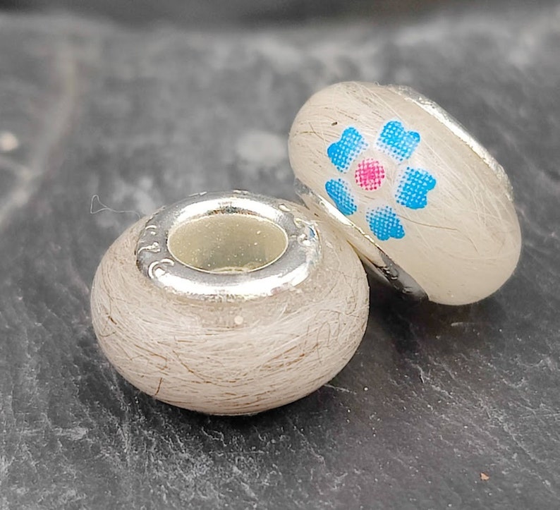 May include: Two decorative beads with silver-colored accents. One bead is round, the other is oval-shaped. The oval bead has a floral design with blue and pink accents. Both beads have a light-colored, textured surface.