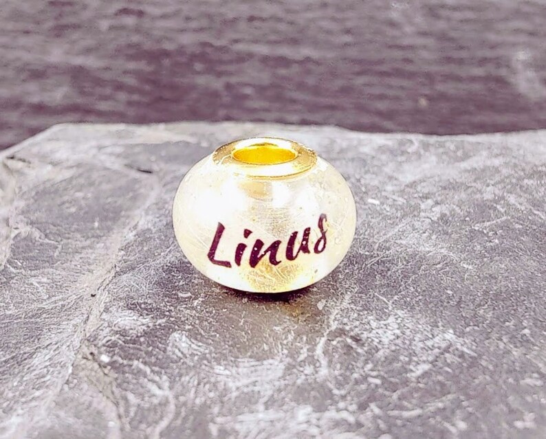 Puede incluir: Una cuenta de color dorado con el nombre "Linus" escrito en una escritura de color rojo oscuro. La cuenta tiene un peque&ntilde;o anillo dorado en la parte superior. El fondo es una superficie oscura y texturizada.