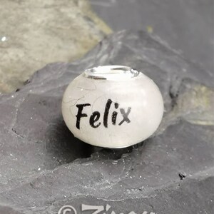 Puede incluir: Una cuenta blanca con el nombre "Felix" en escritura negra. La cuenta tiene una banda de metal plateado alrededor de la parte superior. La cuenta descansa sobre una superficie texturizada de color gris oscuro. El logotipo "Zinou MINIMANUFACTUR" est&aacute; en la parte inferior.