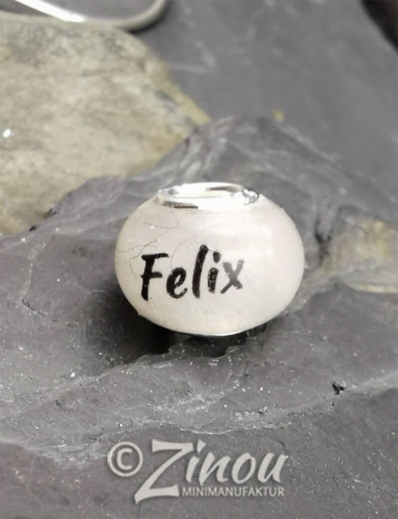 Puede incluir: Una cuenta blanca con el nombre "Felix" en escritura negra. La cuenta tiene una banda de metal plateado alrededor de la parte superior. La cuenta descansa sobre una superficie texturizada de color gris oscuro. El logotipo "Zinou MINIMANUFACTUR" est&aacute; en la parte inferior.