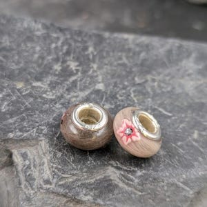 May include: Two wooden beads with silver-colored metal accents. One bead has a pink flower design with a small crystal in the center. The other bead is plain. These beads are likely for jewelry making.