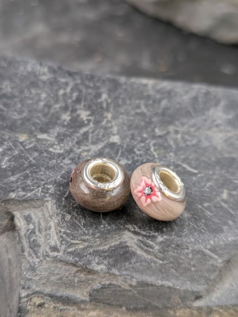 May include: Two wooden beads with silver-colored metal accents. One bead has a pink flower design with a small crystal in the center. The other bead is plain. These beads are likely for jewelry making.