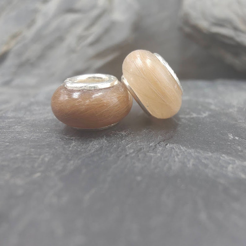 May include: Two round beads with a light brown, textured surface and silver-colored metal accents. One bead is a darker brown color. The beads are likely for jewelry making or crafting.