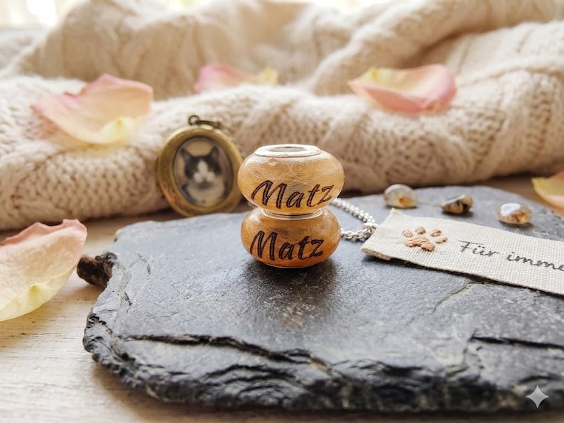 Puede incluir: Un collar con colgante con un peque&ntilde;o medall&oacute;n redondo con una foto de gato y un charm de cuentas de madera de dos partes grabado con el nombre "Matz". El charm est&aacute; conectado a una cadena de plata. Tambi&eacute;n hay una peque&ntilde;a etiqueta con una huella de pata y texto.