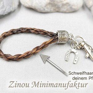 K&ouml;nnte beinhalten: Ein Schl&uuml;sselanh&auml;nger aus geflochtenem braun-schwarzem Pferdehaar mit einer silberfarbenen Metallkappe, einem Karabinerhaken und einem Hufeisenanh&auml;nger mit der Gravur GOOD LUCK. Ein Pfeil zeigt auf das Geflecht mit dem Text Schweifhaare von deinem Pferd.