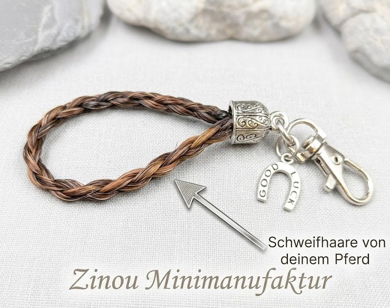 K&ouml;nnte beinhalten: Ein Schl&uuml;sselanh&auml;nger aus geflochtenem braun-schwarzem Pferdehaar mit einer silberfarbenen Metallkappe, einem Karabinerhaken und einem Hufeisenanh&auml;nger mit der Gravur GOOD LUCK. Ein Pfeil zeigt auf das Geflecht mit dem Text Schweifhaare von deinem Pferd.
