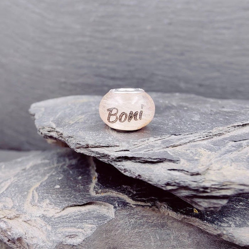 Puede incluir: Una cuenta esf&eacute;rica de color rosa claro con la palabra "Boni" grabada. La cuenta tiene una inserci&oacute;n de metal plateado en la parte superior. La cuenta est&aacute; sobre una superficie texturizada de color gris oscuro.