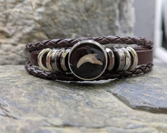 May include: Brown leather bracelet with a braided design and silver-toned metal accents. The focal point is a circular pendant with a black background and a white claw. The bracelet is resting on a textured stone surface.