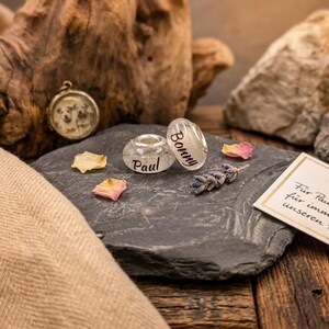 Puede incluir: Una colecci&oacute;n de joyas y recuerdos personalizados. Incluye una cuenta transparente con el nombre "Bonny", una cuenta transparente con el nombre "Paul", un peque&ntilde;o relicario, lavanda seca y una tarjeta con texto. Los art&iacute;culos est&aacute;n dispuestos sobre una piedra gris oscura.