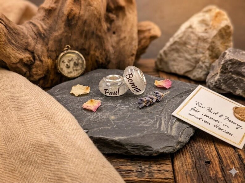 Puede incluir: Una colecci&oacute;n de joyas y recuerdos personalizados. Incluye una cuenta transparente con el nombre "Bonny", una cuenta transparente con el nombre "Paul", un peque&ntilde;o relicario, lavanda seca y una tarjeta con texto. Los art&iacute;culos est&aacute;n dispuestos sobre una piedra gris oscura.