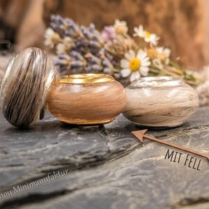 Könnte beinhalten: Drei runde Perlen mit haarartigen Einschlüssen. Die linke Perle ist dunkelbraun und weiß, die mittlere ist hellbraun mit einem goldfarbenen Kern und die rechte ist cremefarben. Ein hölzerner Pfeil zeigt auf sie mit dem Text MIT FELL.