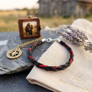 Op de afbeelding: Een gevlochten zwart-rode armband met zilveren sluitingen ligt op een donkergrijze ondergrond, naast een kleine leren fotocase en een gouden kompas hanger. Een linnen zakje en lavendel takjes maken de rustieke scène compleet.