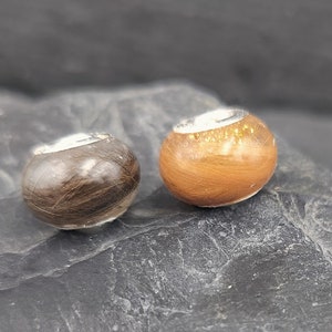 Puede incluir: Dos cuentas redondas con una banda plateada. Una cuenta es marr&oacute;n, la otra es naranja. Las cuentas est&aacute;n hechas de pelo. Las cuentas est&aacute;n sobre una roca gris oscura.