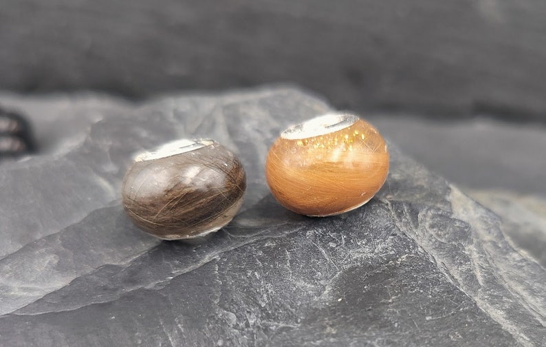 Puede incluir: Dos cuentas redondas con una banda plateada. Una cuenta es marr&oacute;n, la otra es naranja. Las cuentas est&aacute;n hechas de pelo. Las cuentas est&aacute;n sobre una roca gris oscura.