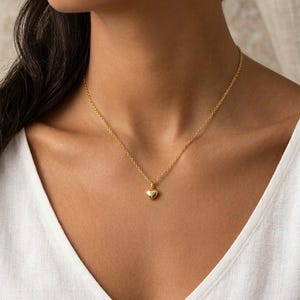 Op de afbeelding: Een goudkleurige ketting met een fijne schakelketting en een kleine, gladde, gepolijste hartvormige hanger. De ketting rust op de hals en toont een eenvoudige, reflecterende metalen afwerking op zowel de kettingschakels als de hartvormige bedel.