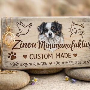Könnte beinhalten: Ein Holzschild mit dem Text "Zinou Minimafaktur CUSTOM MADE" und einem Hundeporträt, einer Katzenillustration und einer Taube. Eine goldene Halskette mit Anhänger befindet sich im Vordergrund. Das Schild liegt auf Steinen.