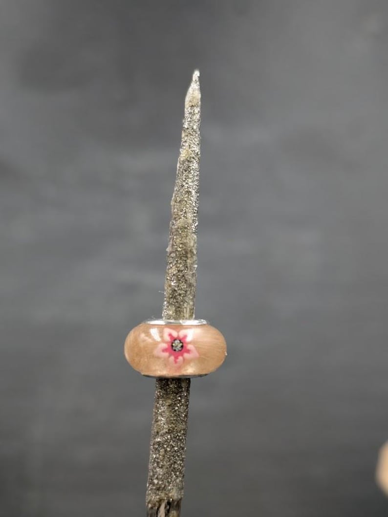 May include: A decorative stick with a light brown bead featuring a pink flower design. The bead is centered on the stick, which is covered in silver glitter. The background is a solid gray.