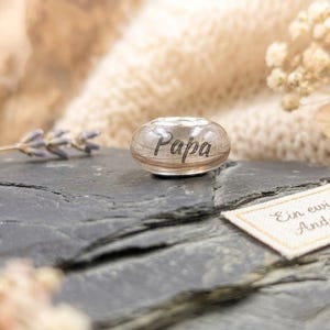 Puede incluir: Una cuenta de vidrio transparente con la palabra "Papa" grabada. Descansa sobre una superficie de piedra gris oscuro, acompa&ntilde;ada de una etiqueta con el texto alem&aacute;n "Ein ewiges Andenken." Flores secas y un fondo suave y texturizado completan la escena.