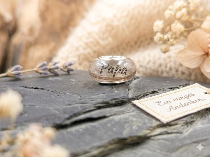 Puede incluir: Una cuenta de vidrio transparente con la palabra "Papa" grabada. Descansa sobre una superficie de piedra gris oscuro, acompa&ntilde;ada de una etiqueta con el texto alem&aacute;n "Ein ewiges Andenken." Flores secas y un fondo suave y texturizado completan la escena.